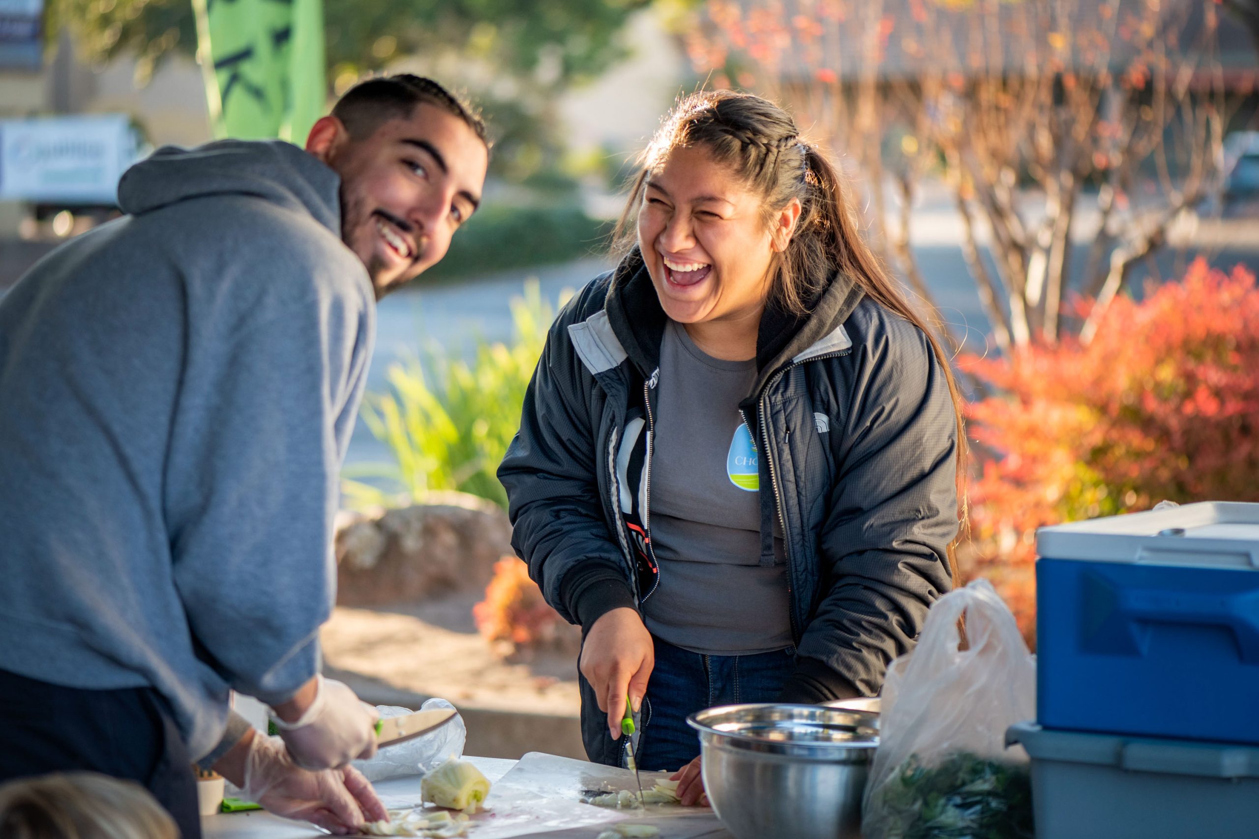 2 CHC staff laughing as they prepare healthy food samples at a local farmers market.