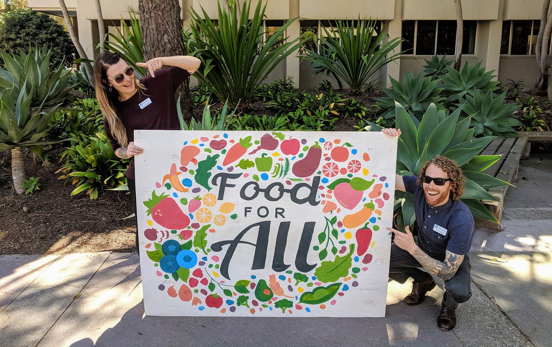 Two people pointing to a sign that says "Food for All"